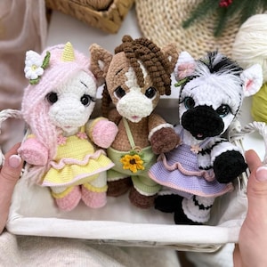 May include: A white basket holds three crocheted animal dolls: a unicorn, a horse, and a zebra. The unicorn has pink hair and a yellow dress. The horse is brown and tan, and the zebra is black and white. Each doll has large eyes and is wearing a dress.