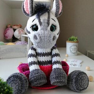 Könnte beinhalten: Ein handgefertigtes gehäkeltes Zebra-Stofftier mit weiß-grau gestreiftem Körper, grauen Hufen und einer rosa-grauen Mähne. Das Zebra hat große, ausdrucksstarke Augen und sitzt auf einer rosa-roten Basis. Zwei graue Kugeln auf beiden Seiten.