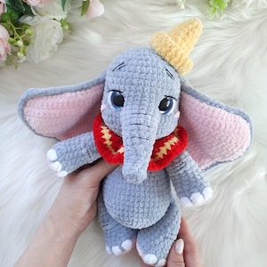 Puede incluir: Un juguete de elefante de ganchillo gris hecho a mano con grandes ojos azules, orejas forradas de rosa y un sombrero amarillo. El elefante tiene un cuello rojo y amarillo y detalles blancos en los pies. El juguete es suave y afelpado.