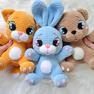 Op de afbeelding: Drie handgemaakte gehaakte knuffels: een oranje kat, een blauw konijn en een bruine beer. Elk pluche speelgoed heeft grote, expressieve ogen met zwarte pupillen, groene of blauwe irissen en roze accenten. De knuffels zijn zacht en knuffelig.