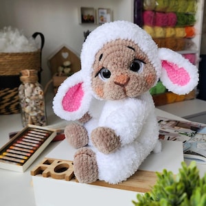 May include: A handmade, plush lamb toy with a white body and head covering. The lamb has a tan face, pink-lined ears, and large, expressive eyes. It is sitting on a white surface, with a wooden accent piece in front.