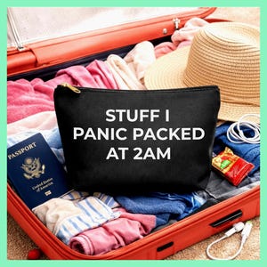 May include: A black zippered pouch with the text "STUFF I PANIC PACKED AT 2AM" sits in an open red suitcase filled with clothes, a straw hat, a passport, and other travel essentials. The pouch has a gold zipper.