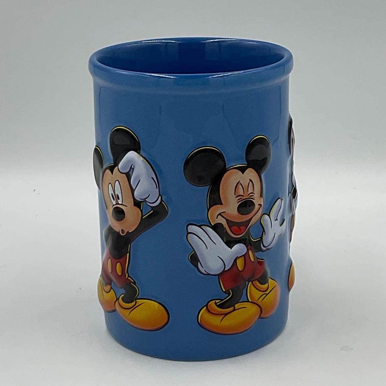 May include: A blue ceramic tumbler featuring Mickey Mouse in various poses. The tumbler has a cylindrical shape with a smooth, glossy finish. Mickey Mouse is depicted in his classic outfit with red shorts, yellow shoes, and white gloves.