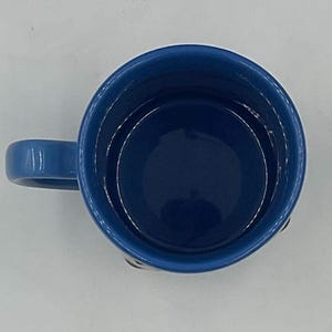 May include: A top-down view of a blue ceramic mug with a matching handle. The interior of the mug is a darker shade of blue. The mug is set against a white background.
