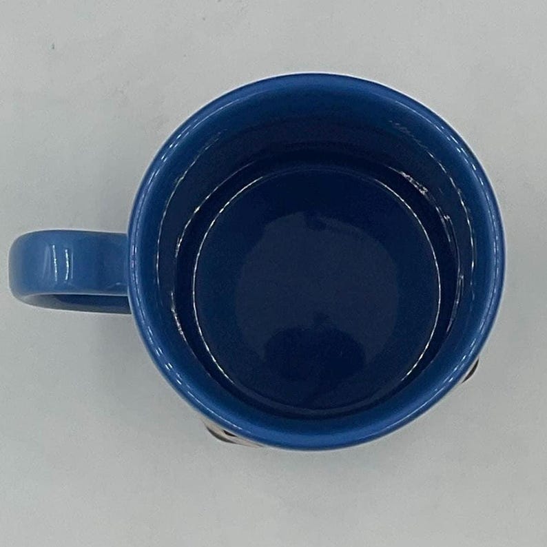 May include: A top-down view of a blue ceramic mug with a matching handle. The interior of the mug is a darker shade of blue. The mug is set against a white background.