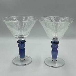 Puede incluir: Dos copas de martini de cristal transparente con cuencos anchos y acampanados que presentan una textura de burbujas. Cada copa se apoya en un tallo sólido de color azul intenso con un diseño redondeado y escalonado y una base circular.