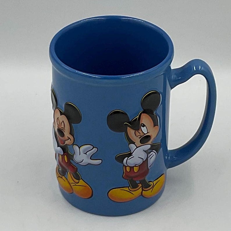 May include: A blue ceramic mug featuring two images of Mickey Mouse. The mug has a curved handle and a glossy finish. Mickey Mouse is depicted in his classic outfit with red shorts, yellow shoes, and white gloves.