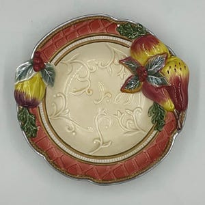 May include: A round ceramic plate featuring a cream-colored center with embossed scrollwork patterns. The rim has a textured, reddish-orange lattice design. Three-dimensional ceramic pears and green leaves are attached to the sides of the plate.