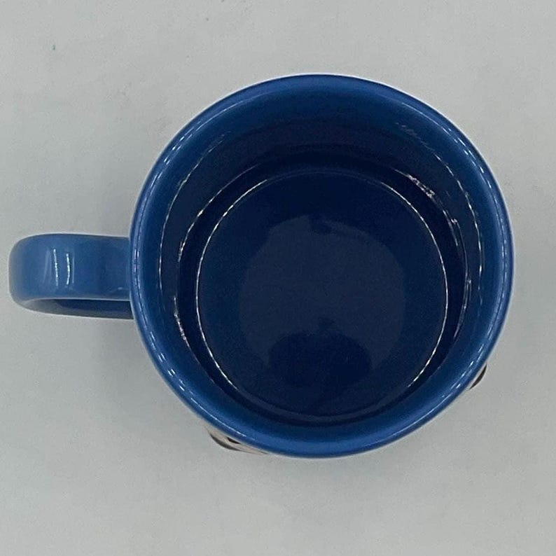May include: A top-down view of a ceramic mug featuring a deep blue interior and a matching blue handle. The rim of the mug is circular, and the smooth, glossy finish of the blue glaze is visible throughout the inside and on the exterior handle.