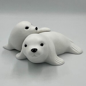 Peut inclure: Une paire de figurines de phoque en céramique blanche au fini mat. Un phoque est placé au premier plan face à l'avant, tandis que le second est légèrement en retrait et incliné sur le côté. Les deux figurines ont des yeux et des nez noirs.