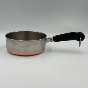 Op de afbeelding: Een roestvrijstalen steelpan met een koperkleurige bodem en een zwarte handgreep. De handgreep is voorzien van een zilverkleurig metalen bevestigingspunt en een kleine ronde metalen ring aan het uiteinde om de pan op te hangen.