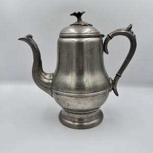 May include: A silver-colored metal teapot with a curved spout and handle. The lid is topped with a decorative floral element. The teapot has a bulbous body and sits on a round base. A classic piece for serving tea or coffee.