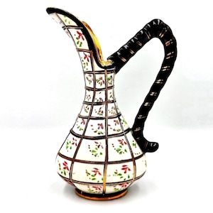May include: A ceramic pitcher featuring a white base with a black grid pattern and small red and green floral accents. The pitcher has a tall, slender neck, a wide base, and a dark, twisted handle with gold-toned detailing along the edges.