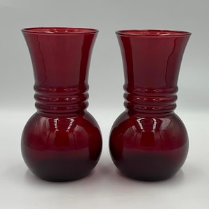 May include: A pair of matching deep red glass vases, each featuring a rounded bulbous base that tapers into a slender neck with three horizontal ribbed rings encircling the middle section and a flared opening at the top rim.