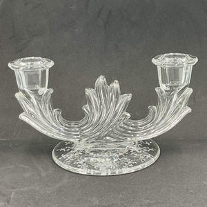 May include: A clear glass double candle holder featuring two cylindrical candle cups connected by a central decorative leaf-like structure. The base is circular and features an etched floral pattern across its surface, providing a stable foundation.