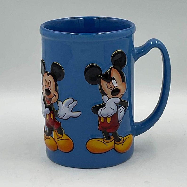 May include: Blue ceramic mug featuring two images of Mickey Mouse. Mickey is depicted in his classic outfit with red shorts, yellow shoes, white gloves, and a black head. The mug has a large handle and a smooth, glossy finish.