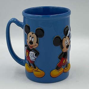 May include: A blue ceramic mug featuring embossed images of Mickey Mouse. The mug has a curved handle and a glossy finish. Mickey is depicted in his classic outfit with red shorts, yellow shoes, and white gloves.