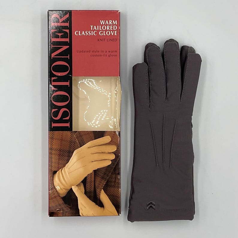 May include: A pair of dark gray Isotoner gloves with three vertical stitched lines on the back of the hand and a small chevron logo near the wrist, presented next to a vertical product box labeled Isotoner Warm Tailored Classic Glove.