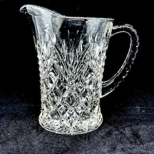 May include: Clear glass pitcher with a diamond pattern and a decorative handle. The pitcher has a spout for easy pouring. The glass is thick and has a textured surface. The pitcher is likely used for serving beverages.