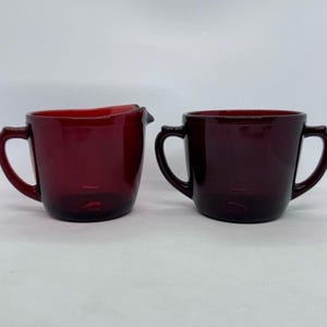 May include: A deep red glass creamer jug with a single handle and a pouring spout, paired with a matching deep red glass sugar bowl featuring two side handles. Both pieces are made of smooth, translucent dark red glass with a glossy finish.