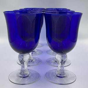 May include: Eight cobalt blue wine glasses are displayed. Each glass has a rounded bowl, a textured stem, and a clear, circular base. The glasses are arranged in two rows, highlighting their elegant design.