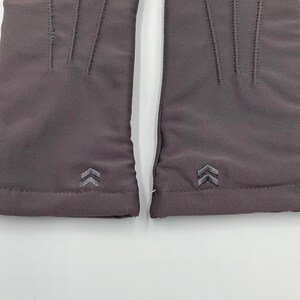 May include: A pair of dark gray gloves featuring three vertical stitched lines on the back of the hand and a small, embroidered double-chevron logo near the wrist cuff. The fabric has a smooth, matte finish and a clean, minimalist construction.