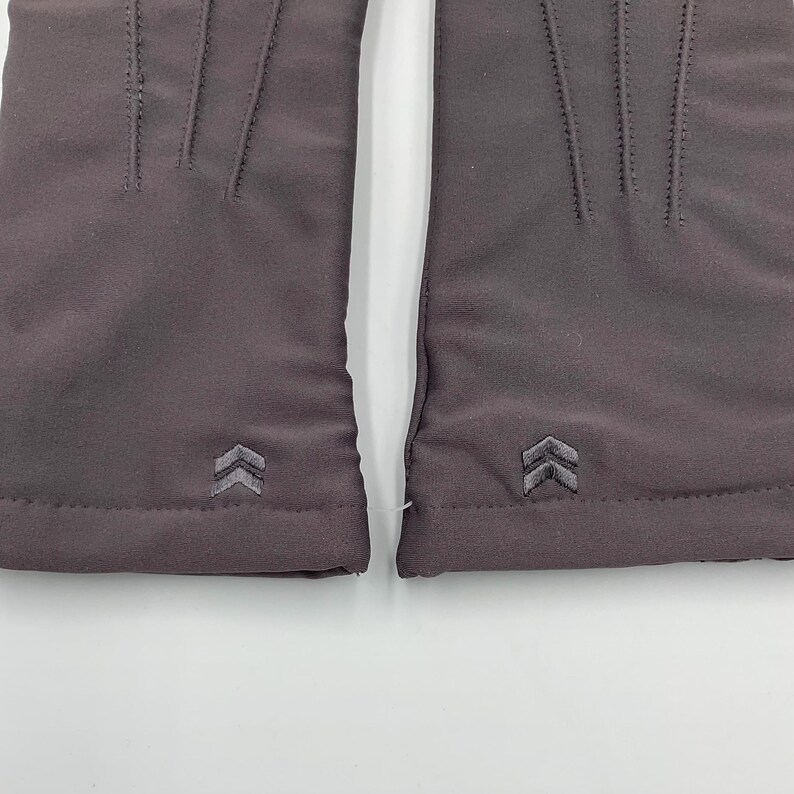May include: A pair of dark gray gloves featuring three vertical stitched lines on the back of the hand and a small, embroidered double-chevron logo near the wrist cuff. The fabric has a smooth, matte finish and a clean, minimalist construction.