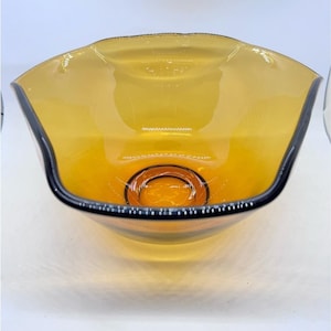 May include: An amber-coloured glass bowl with a black rim. The bowl has a unique, wavy shape and a circular indentation at the base. The glass is transparent, allowing light to pass through, and the bowl appears to be empty.
