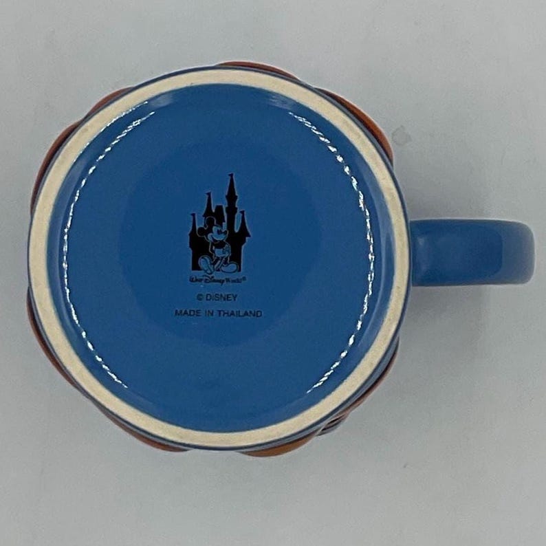 May include: Blue ceramic mug with a handle, featuring a Mickey Mouse and castle design. The mug has a woven brown and orange pattern around the rim. The bottom of the mug has the text "Walt Disney World", "&copy; Disney", and "MADE IN THAILAND".