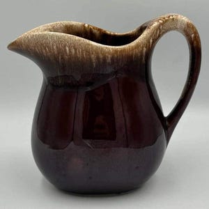 Può includere: Una brocca vintage in ceramica marrone con corpo arrotondato e manico curvo. Il bordo superiore e il manico presentano una glassa strutturata marrone chiaro, in contrasto con il corpo marrone scuro lucido. La brocca ha un beccuccio.