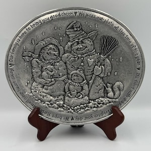 Puede incluir: Un plato decorativo de color peltre con una familia de muñecos de nieve. El plato tiene un diseño en relieve con una familia de muñecos de nieve, una ardilla y texto alrededor del borde. El plato se exhibe en un soporte de madera.