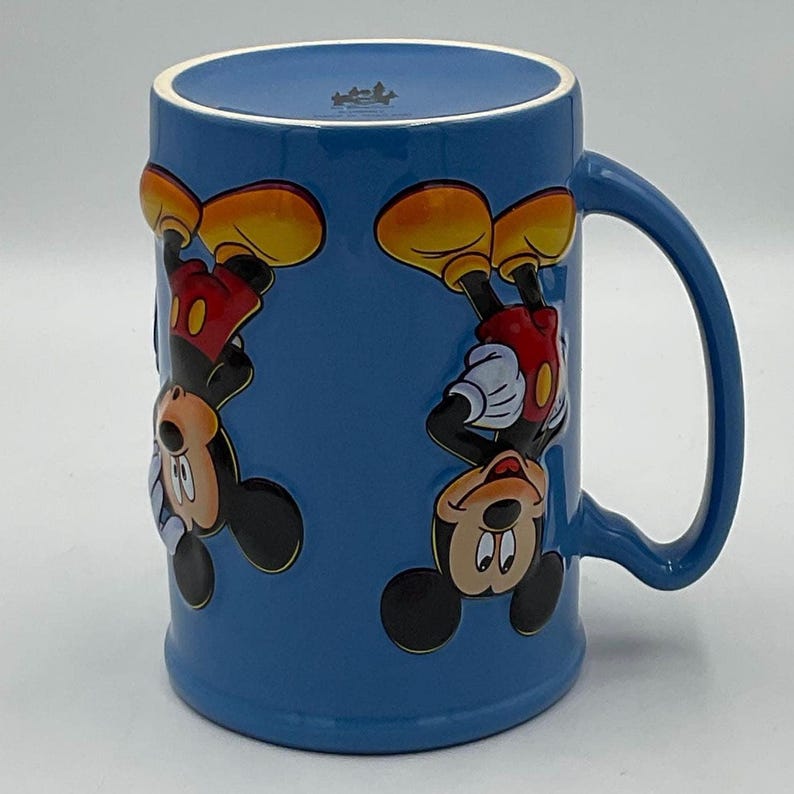 May include: Blue ceramic mug featuring Mickey Mouse in a repeating pattern. Mickey is depicted upside down with his signature yellow shoes, red shorts, white gloves, and black face. The mug has a curved handle.