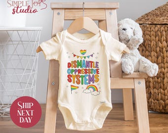 May include: A white baby onesie with a rainbow-colored graphic that says "Dismantle Oppressive Systems". The graphic also includes a rainbow, a heart, and clouds.