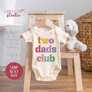 May include: A white baby onesie with the text "two dads club" in rainbow colors. The onesie is hanging on a wooden hanger.