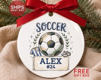 May include: A round white ceramic ornament featuring a central soccer ball graphic, a blue and white jersey, and a pair of cleats. The text reads SOCCER, ALEX, and #24. A red satin ribbon is tied in a bow at the top of the ornament.