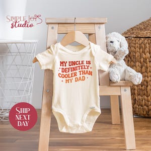 May include: Cream-colored baby bodysuit with the text "MY UNCLE IS + DEFINITELY + COOLER THAN + MY DAD +" in red. The bodysuit is hanging on a wooden hanger. A white stuffed dog sits on a small wooden chair.