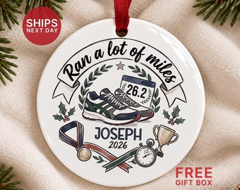 May include: A round white ceramic ornament with a red ribbon, featuring a graphic of running shoes, a marathon bib labeled 26.2, a medal, a trophy, and a stopwatch. The text reads Ran a lot of miles, JOSEPH, and 2026, surrounded by laurel leaves.