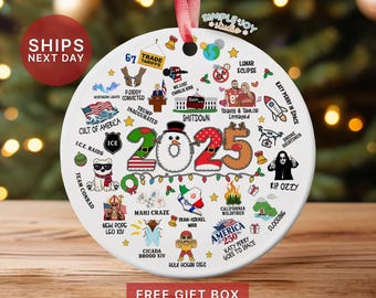 May include: A white ceramic Christmas ornament with a red ribbon. The ornament features the year "2025" with various illustrations and text, including "Shutdown", "Katy Perry in Space", and "America 250".