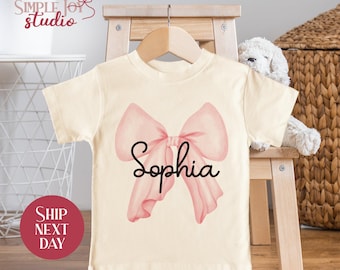 May include: A cream colored t-shirt with a pink bow and the name "Sophia" written in black cursive.