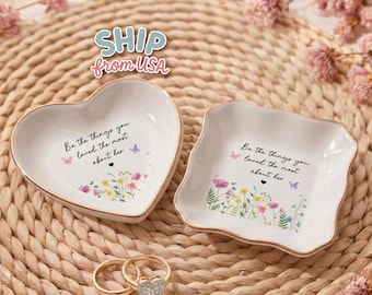 May include: Two white ceramic trinket dishes, one heart-shaped and one square with scalloped edges, both featuring the text "Be the things you loved the most about her" with colorful floral and butterfly illustrations, plus two gold-tone rings.