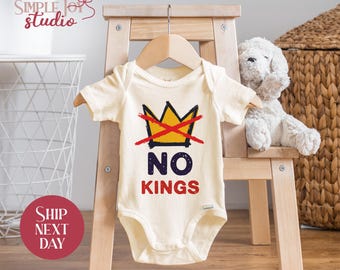May include: Cream-colored baby bodysuit with a graphic of a yellow crown crossed out with a red line, and the words "NO KINGS" in navy and red. The bodysuit is hanging on a wooden hanger on a small wooden chair.