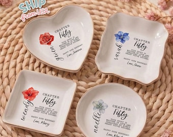 May include: Four white ceramic trinket dishes in heart, square, and round shapes. Each dish features a floral illustration, the text 'CHAPTER FIFTY', a personalized name, and the message 'YOUR STORY IS WRITTEN IN LOVE AND TREASURED BY EVERY HEART YOU'VE TOUCHED. HAPPY 50TH BIRTHDAY. LOVE, [NAME]'.