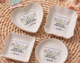 May include: Four white ceramic trinket dishes in heart, square, round, and scalloped shapes. Each dish has a gold rim and features a floral design with the text "SPEECH THERAPIST" and a name. The text "NEVER FORGET THE DIFFERENCE THAT YOU MAKE" is also included.