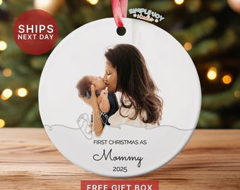May include: A white ceramic Christmas ornament with a photo of a mother kissing a baby. The ornament has a red ribbon for hanging and the text "FIRST CHRISTMAS AS Mommy 2025".