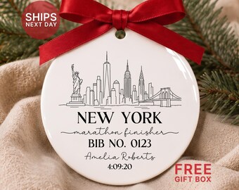 May include: A round white ceramic ornament featuring a black line drawing of the New York City skyline, including the Statue of Liberty and Brooklyn Bridge. Text reads: NEW YORK, marathon finisher, BIB NO. 0123, Amelia Roberts, 4:09:20. A red ribbon is tied at the top.