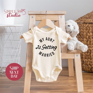 May include: Cream-colored baby onesie with the text "MY AUNT Is Getting MARRIED" in black. The onesie is hanging on a wooden hanger, displayed on a small wooden chair. A stuffed white dog sits next to it.