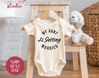 May include: Cream-colored baby onesie with the text "MY AUNT Is Getting MARRIED" in black. The onesie is hanging on a wooden hanger, displayed on a small wooden chair. A stuffed white dog sits next to it.