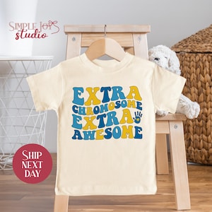 May include: A white short-sleeved t-shirt with the text "EXTRA CHROMOSOME EXTRA AWESOME" in blue and yellow letters. The text is in a retro font style.