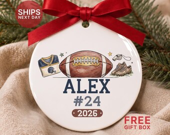 May include: A round white ceramic ornament featuring a red satin ribbon bow at the top. The design includes a football, a jersey, a whistle, and cleats. The text reads ALEX, #24, and 2026 in dark blue and brown lettering.
