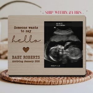 May include: A wooden picture frame with the text "Someone wants to say hello" and "BABY ROBERTS Arriving January 2026" engraved on it. The frame holds a sonogram image. The frame is light brown.